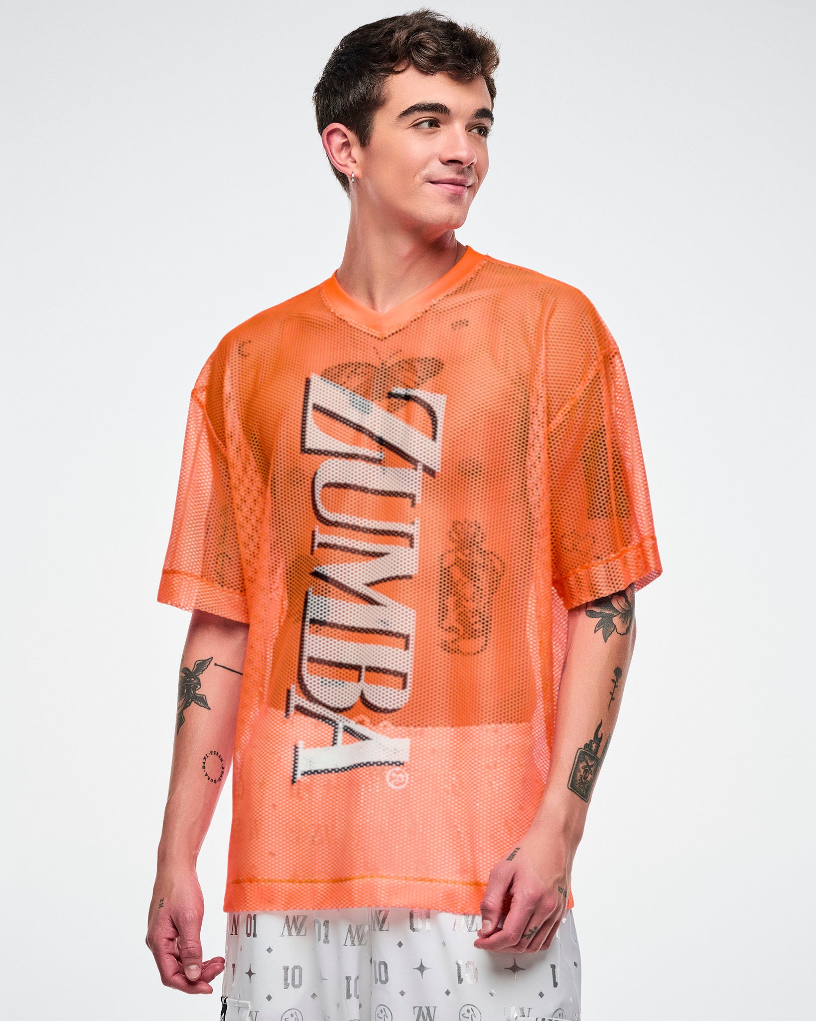 Male model wearing an oversized orange mesh Zumba top with bold vertical “ZUMBA” silver lettering paired with white and silver pants.