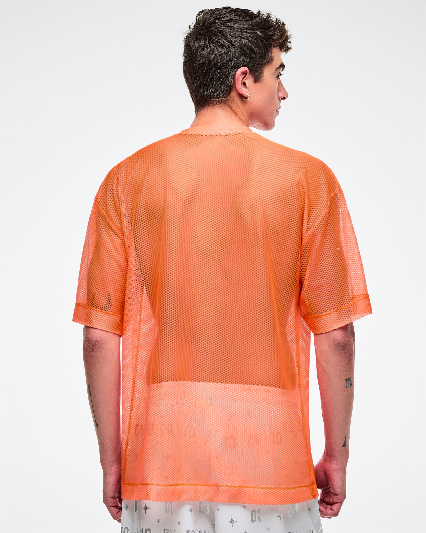 Back view of a male model wearing an oversized orange mesh Zumba top with bold vertical “ZUMBA” silver lettering paired with white and silver pants.