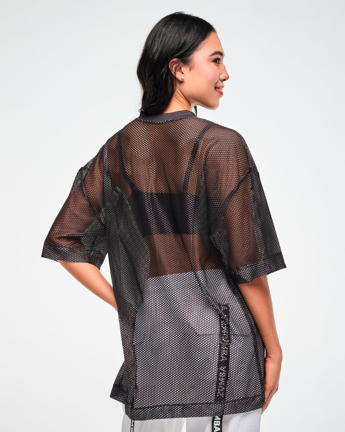 Back view of the black mesh oversized top, female model, showing the sheer mesh layered over a black bra and white pants.