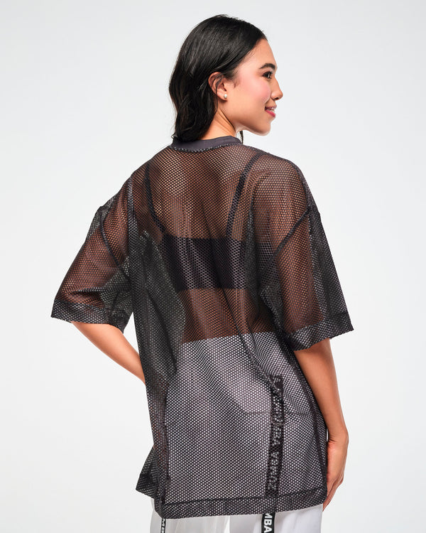 Back view of the black mesh oversized top, female model, showing the sheer mesh layered over a black bra and white pants.