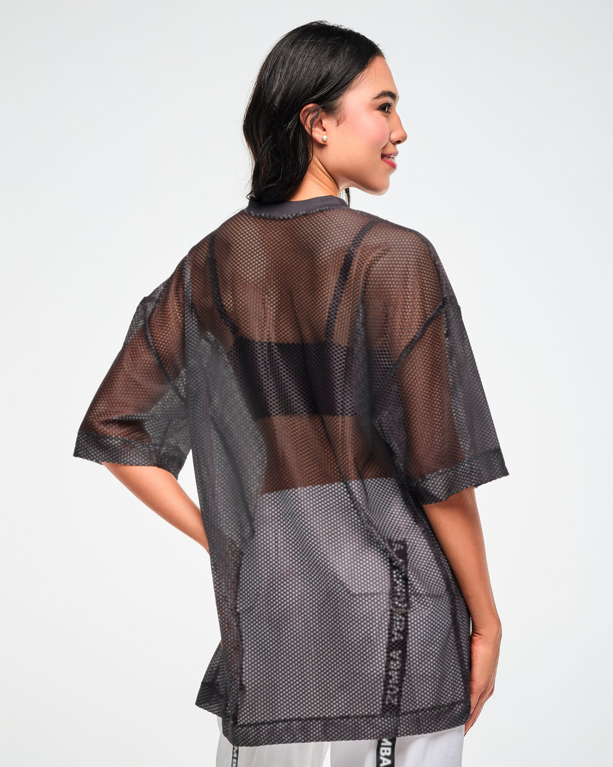 Back view of the black mesh oversized top, female model, showing the sheer mesh layered over a black bra and white pants.