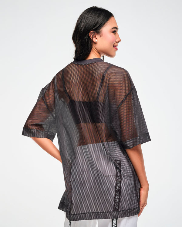 Back view of the black mesh oversized top, female model, showing the sheer mesh layered over a black bra and white pants.