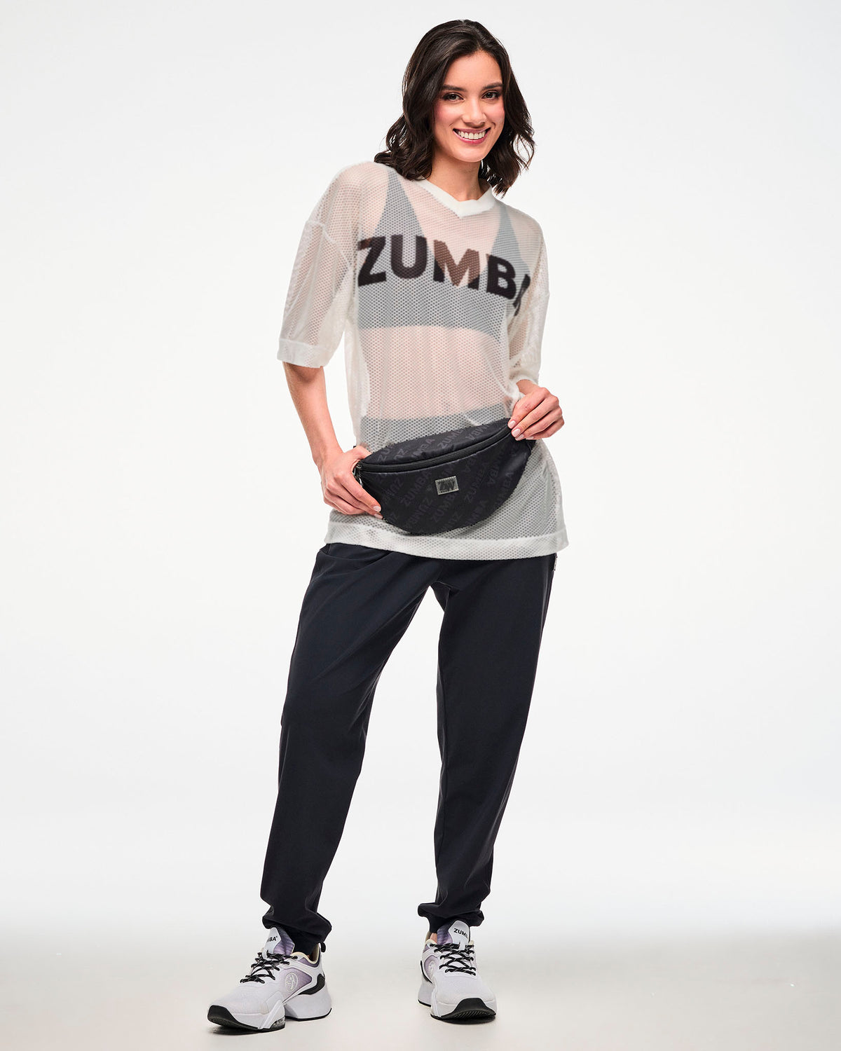 White mesh oversized top with a black 'ZUMBA' logo over a black bra, styled with black track pants and a black 'ZUMBA'-print crossbody bag, female model.