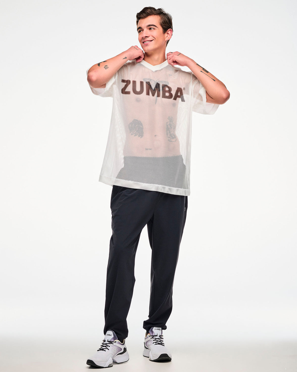 Male model in a white mesh oversized top with a black 'ZUMBA' logo, tugging the collar with both hands, paired with black track pants and white shoes.