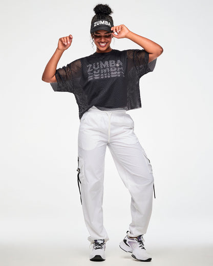 Black mesh cropped top over a black t-shirt with white 'ZUMBA' text, worn by a female model with a black 'ZUMBA' visor white cargo pants and white shoes.