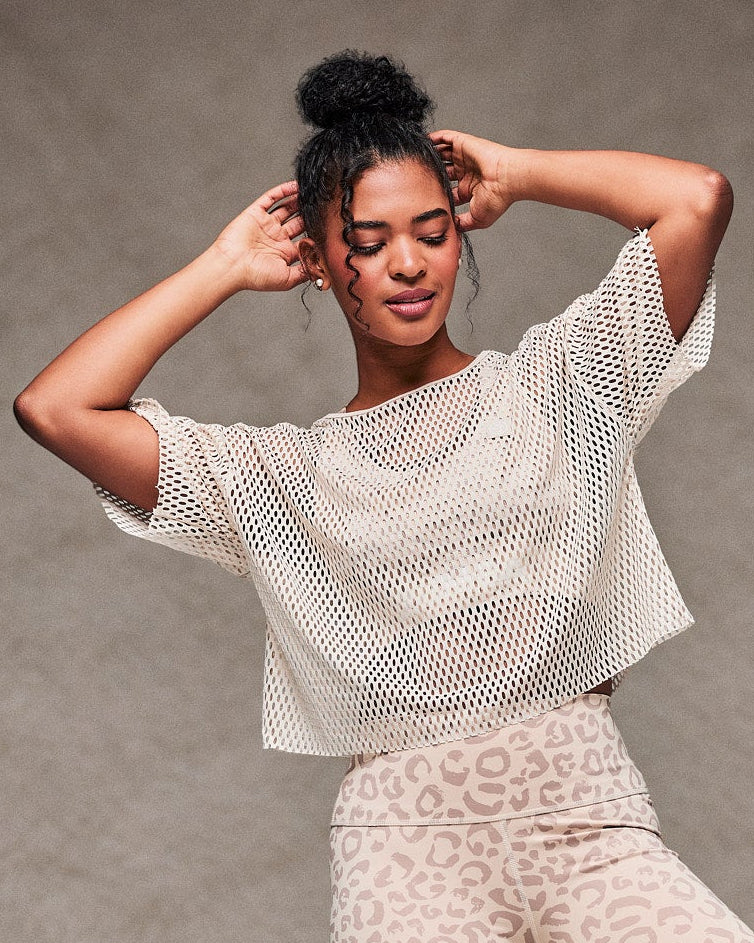 Female model mid-dance with both hands at her hair in the beige mesh crop top, paired with beige leopard print biker shorts and beige shoes. Taupe faux fur backdrop.