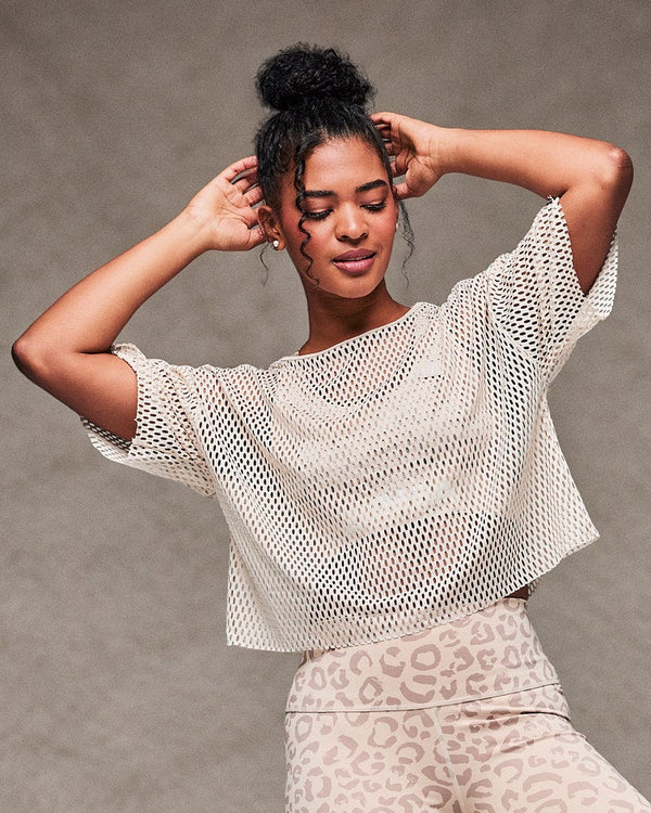 Female model mid-dance with both hands at her hair in the beige mesh crop top, paired with beige leopard print biker shorts and beige shoes. Taupe faux fur backdrop.