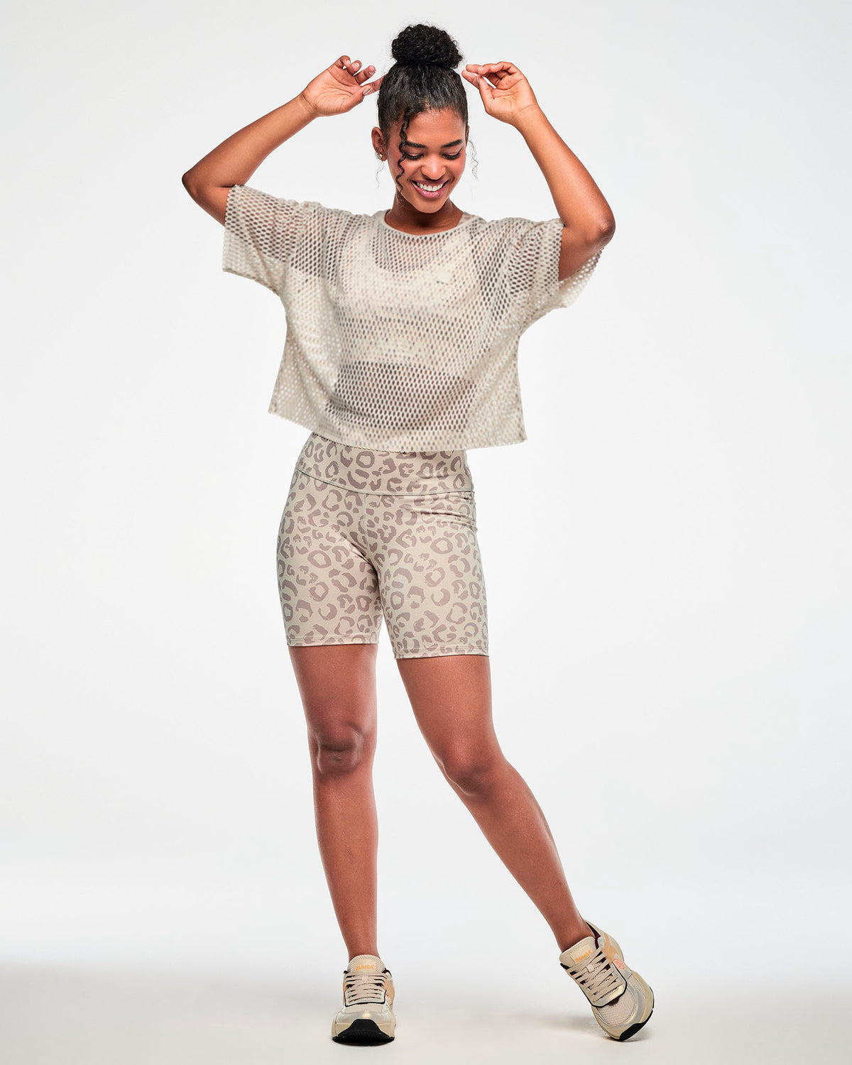 Full view of a female model with hands above her head, wearing a beige open-hole mesh crop top over a beige bra, paired with beige leopard print biker shorts and beige shoes.