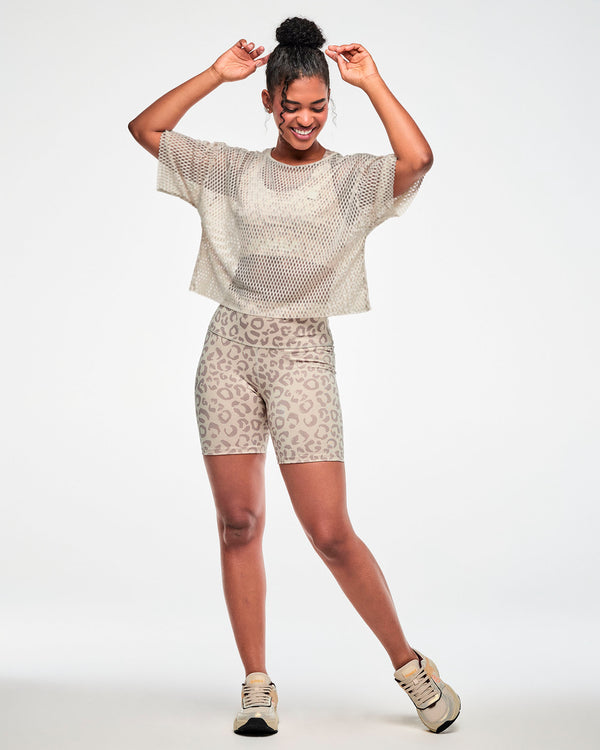Full view of a female model with hands above her head, wearing a beige open-hole mesh crop top over a beige bra, paired with beige leopard print biker shorts and beige shoes.