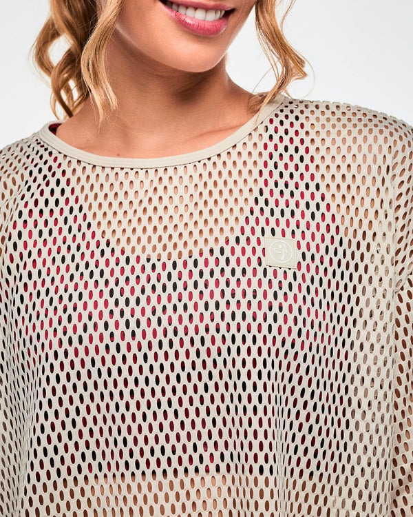Close-up of a beige open-hole mesh crop top, showing the bound crew neckline and a square beige patch with a white Zumbito logo on the upper left chest, with a dark red leopard print bra visible underneath.