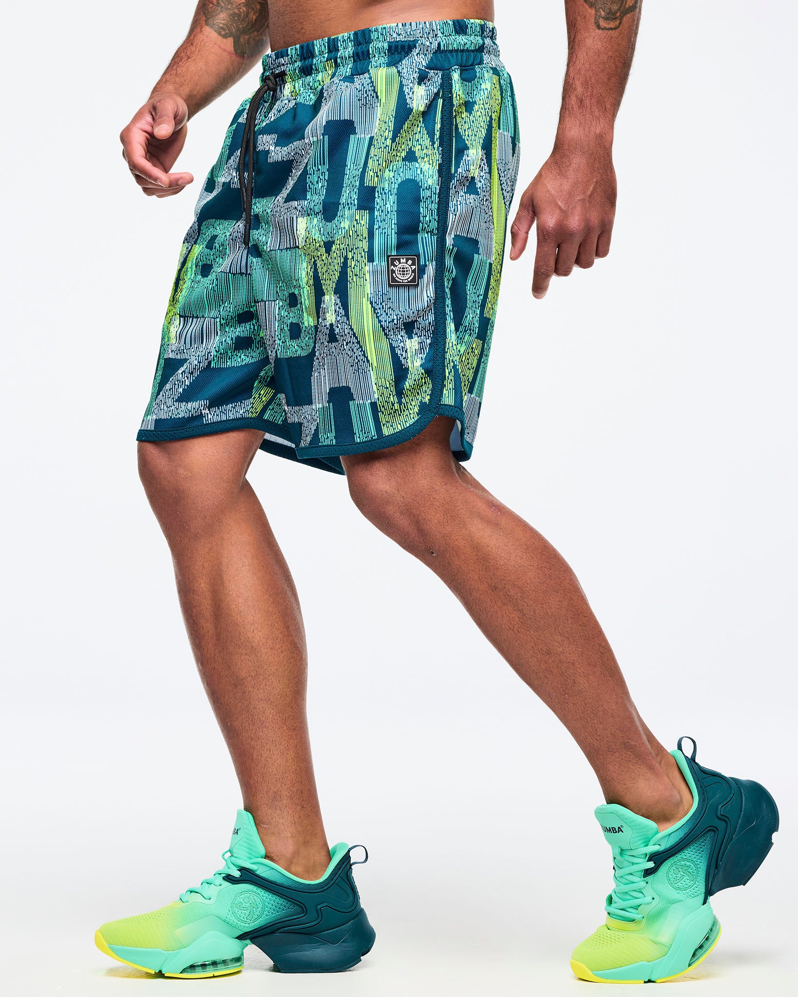 Side view of the teal-and-lime all-over print athletic shorts on a male model, showing the logo patch near the hem and curved side slit, paired with aqua-to-lime sneakers.