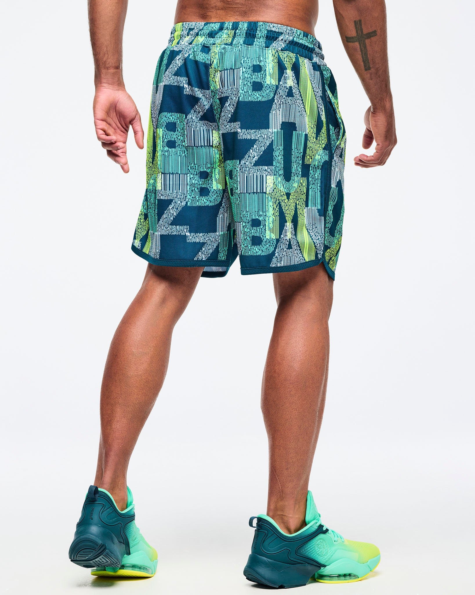Back view of the teal-and-lime all-over print athletic shorts on a male model, showing the elastic waistband and curved hem, paired with aqua-to-lime sneakers.