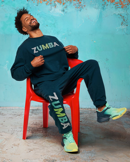 Teal jogger sweatpants with vertical “ZUMBA” leg graphic, styled with a matching teal sweatshirt, modeled by a male sitting on a red chair against a turquoise wall and wearing teal-to-lime athletic sneakers.