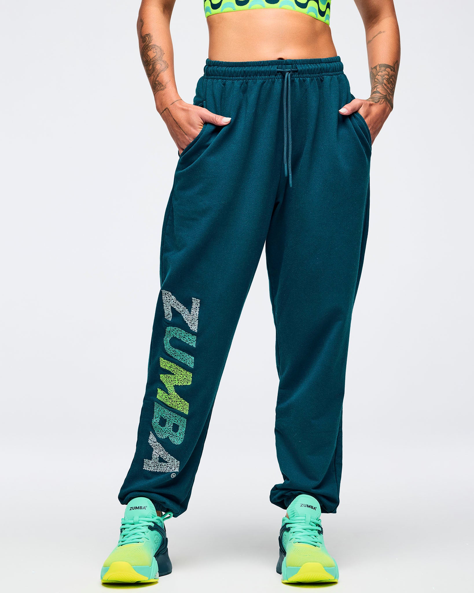 Teal jogger sweatpants with elastic drawstring waistband and relaxed tapered fit, modeled by a female wearing ombre green to lime athletic sneakers on a white studio background.