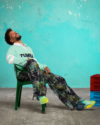 Lifestyle shot of a male model lounging on a green chair wearing the black wide-leg printed track pants, mint-and-white collared sweatshirt with “ZUMBA” across the chest, and aqua-to-lime sneakers.