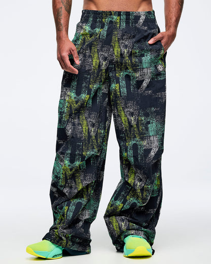 Front view of the black wide-leg printed track pants on a male model with hands in pockets, showing the abstract teal-and-lime pattern and loose legs, paired with aqua-to-lime sneakers.