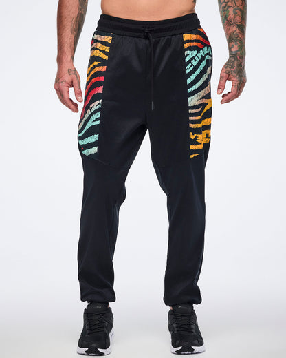 Male model wearing black jogger pants with multicolor zebra-print paneling on the upper legs, a drawstring waistband, tapered legs, and black sneakers with white soles.