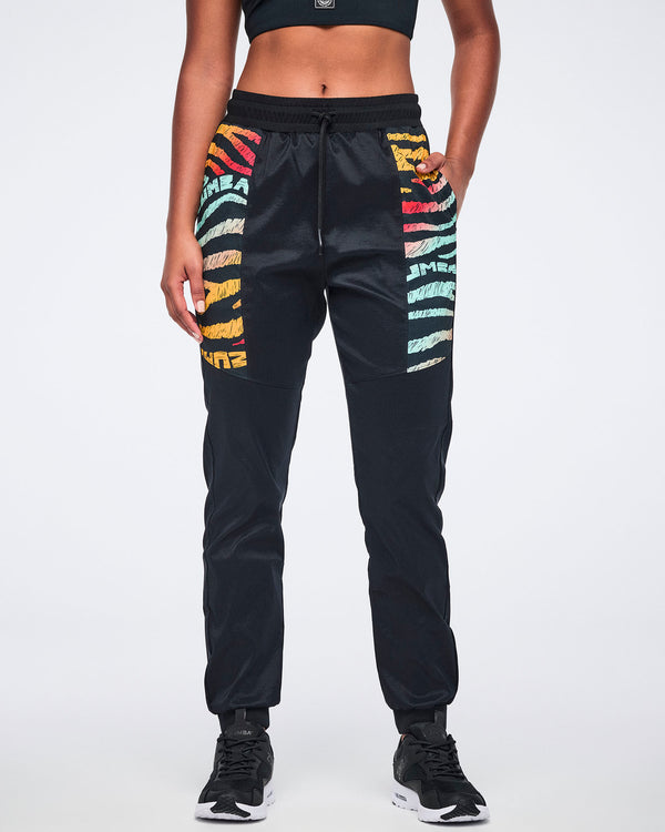 Female model wearing black jogger pants with multicolor zebra-print paneling on the upper legs, a drawstring waistband, tapered legs, and black sneakers with white soles.