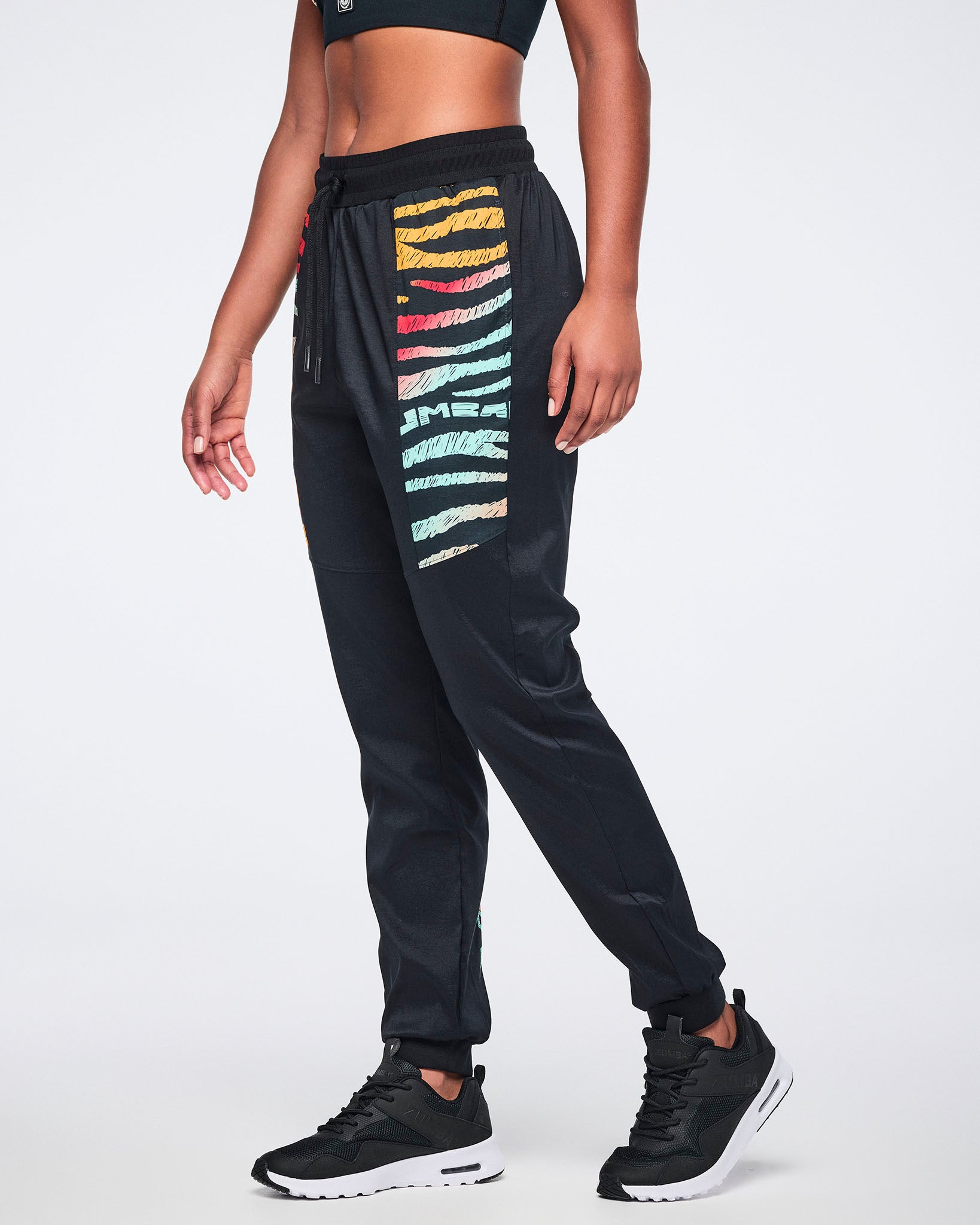 Female model wearing black jogger pants with multicolor zebra-print paneling on the upper legs, a drawstring waistband, tapered legs, and black sneakers with white soles.