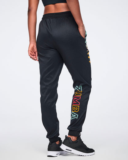 Female model wearing black jogger pants shown from the back with a relaxed fit, multicolor Zumba logo on the lower leg, tapered cuffs, and black sneakers with white soles.