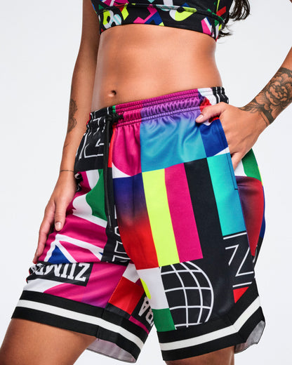 Close-up studio shot of a female model wearing vibrant, geometric-patterned shorts, her left hand tucked into the pocket, set against a white background.