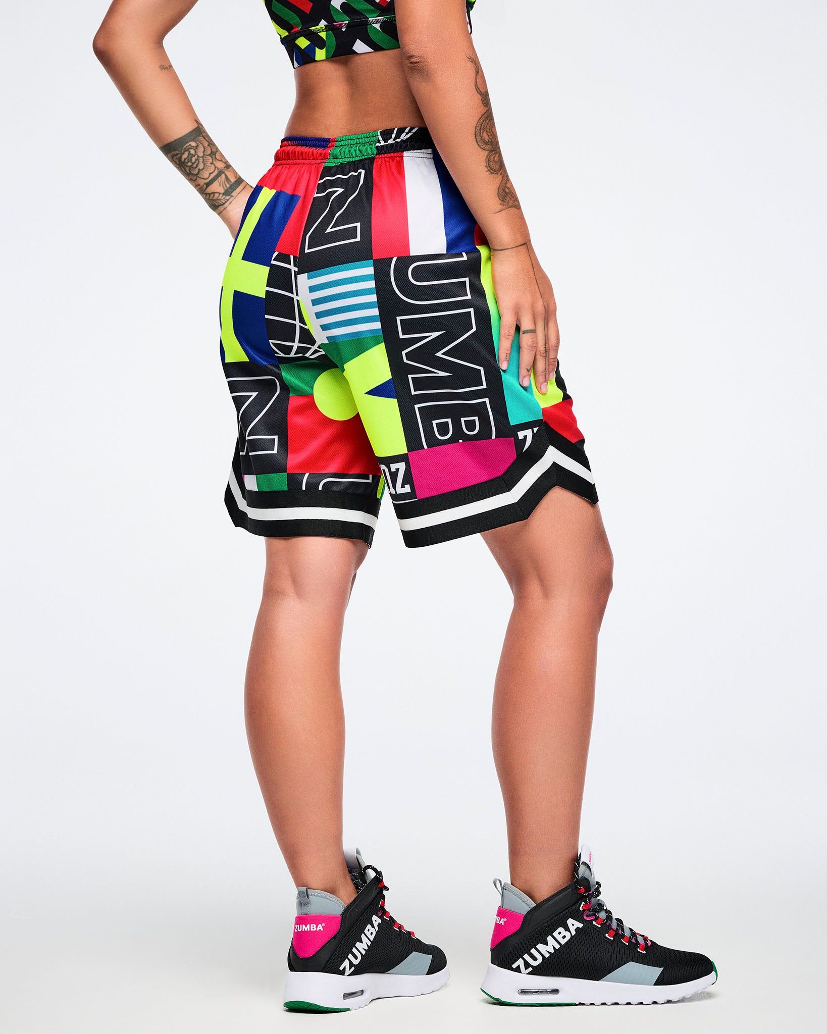 Female model from the back in bold multicolor graphic basketball shorts, black-pink high-tops against a white studio background.