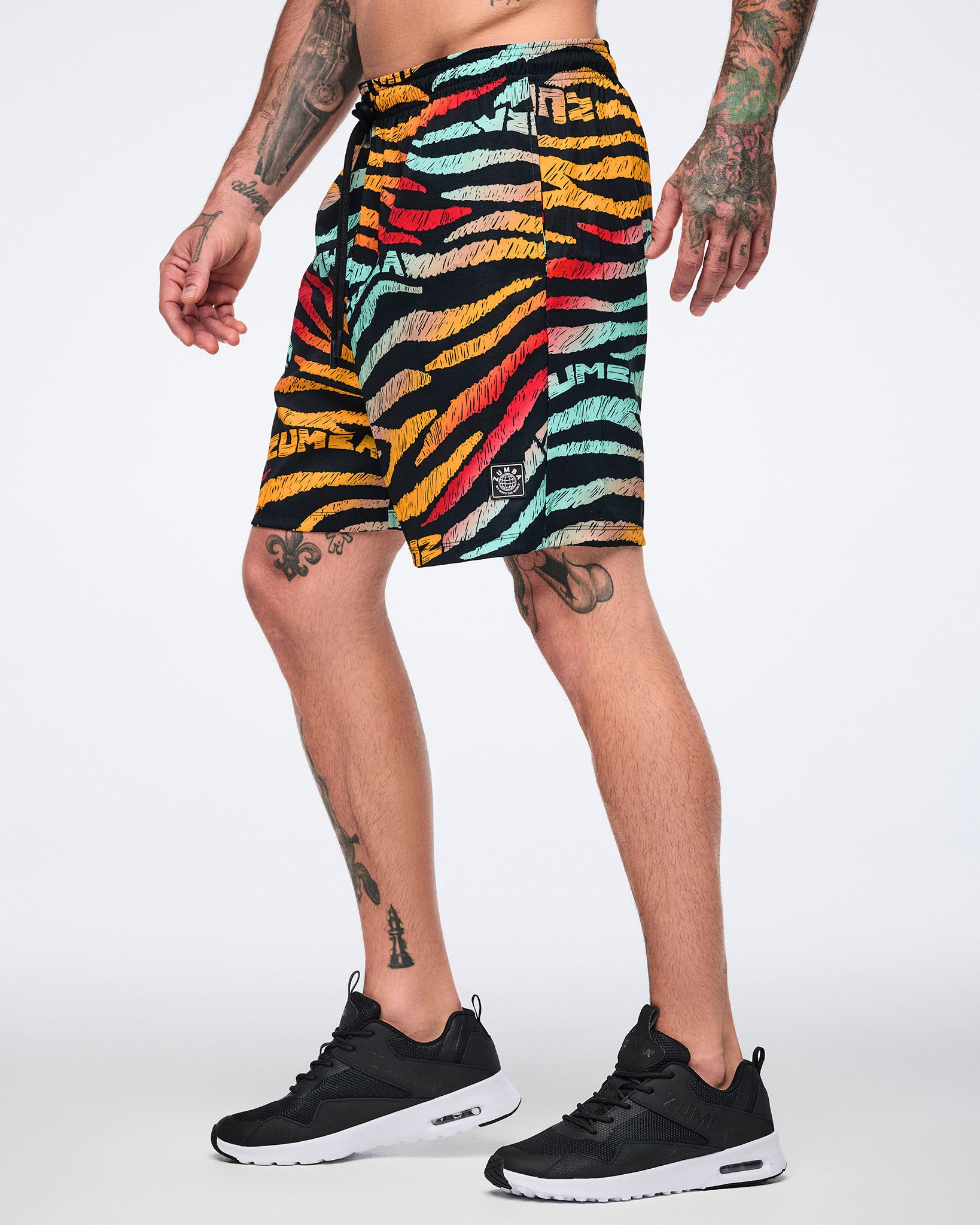 Male model wearing knee-length athletic shorts in a black zebra-stripe print with multicolor brushstroke stripes in yellow, orange, red, mint, and beige, shown in side view with a black drawstring waistband and black low-top sneakers with white soles.