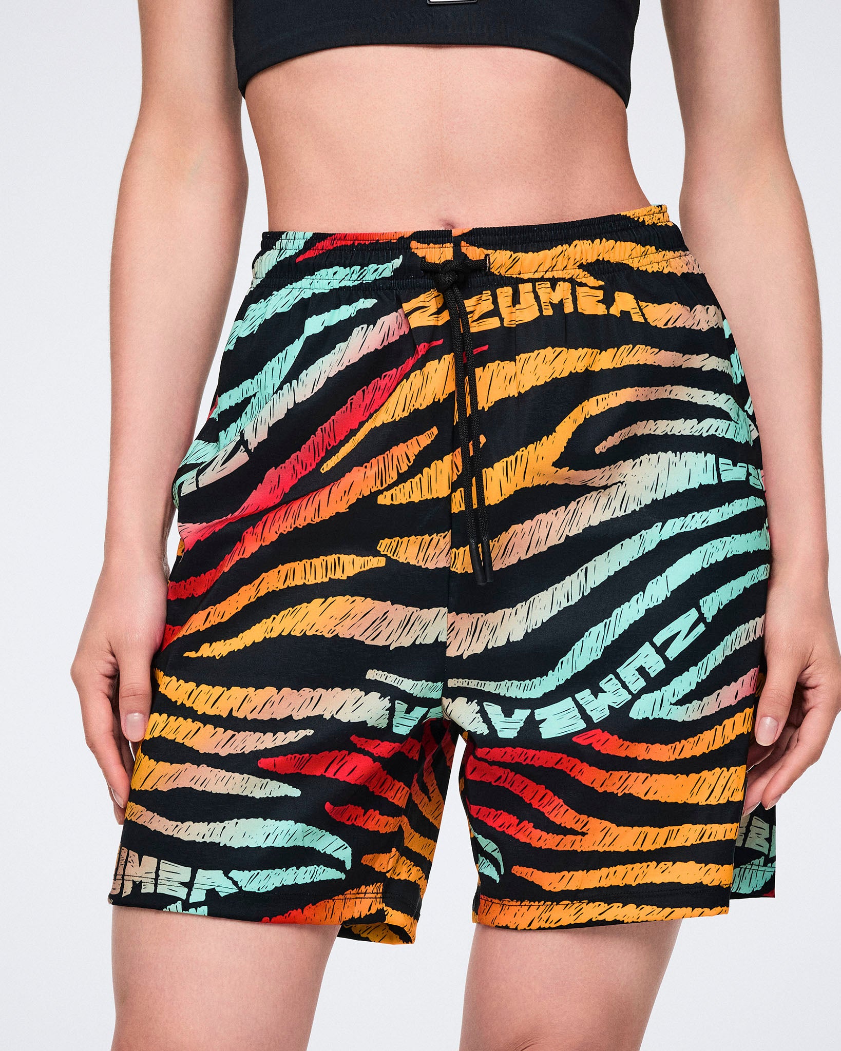 Female model wearing knee-length athletic shorts in a black zebra-stripe print with multicolor brushstroke stripes in yellow, orange, red, mint, and beige, featuring a black drawstring waistband and a relaxed fit.