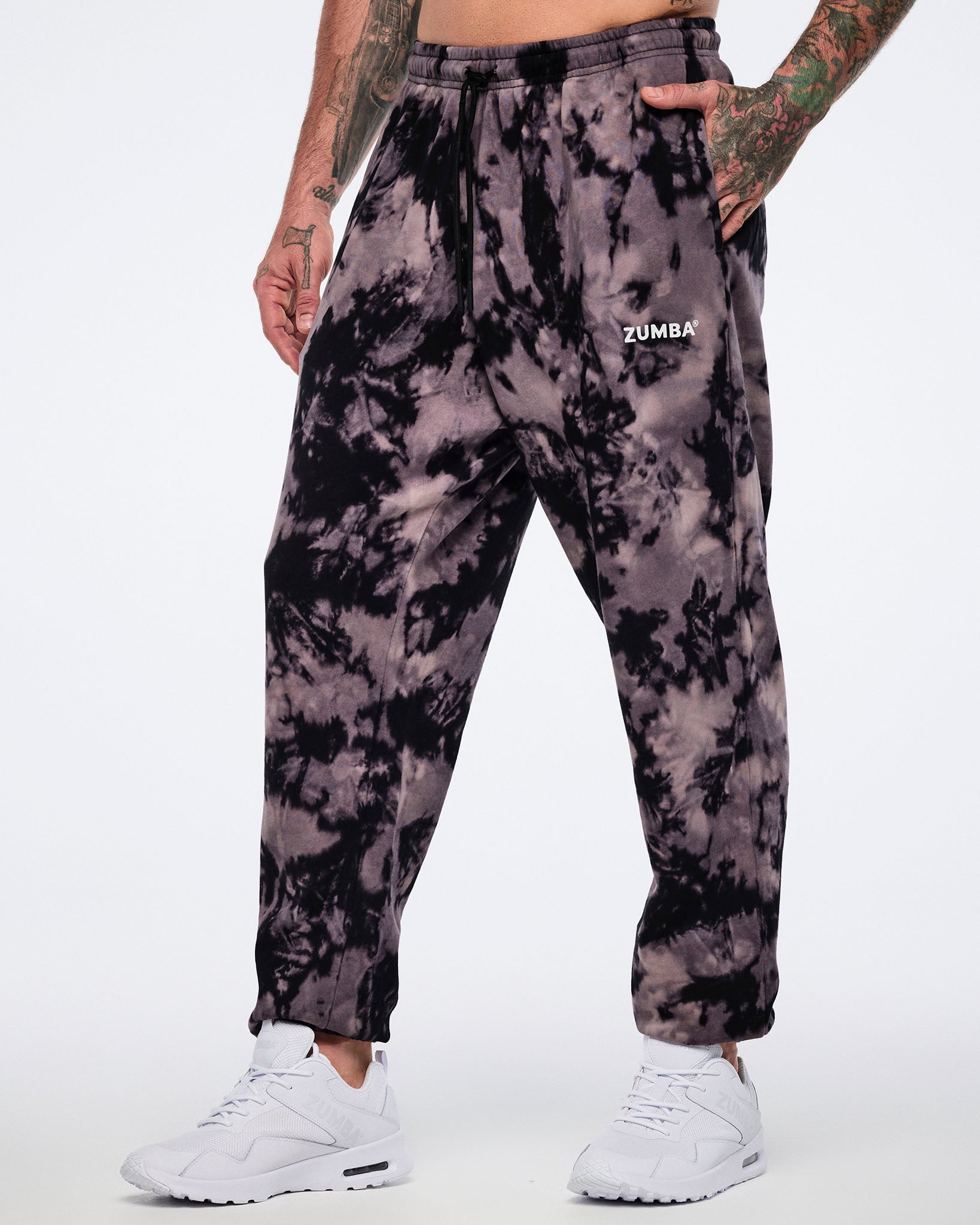 Black and tan tie-dye sweatpants with a small white Zumba logo on the front. Male model wears the relaxed-fit sweatpants with a black drawstring waistband, side pockets, tapered legs, and white low-top sneakers.