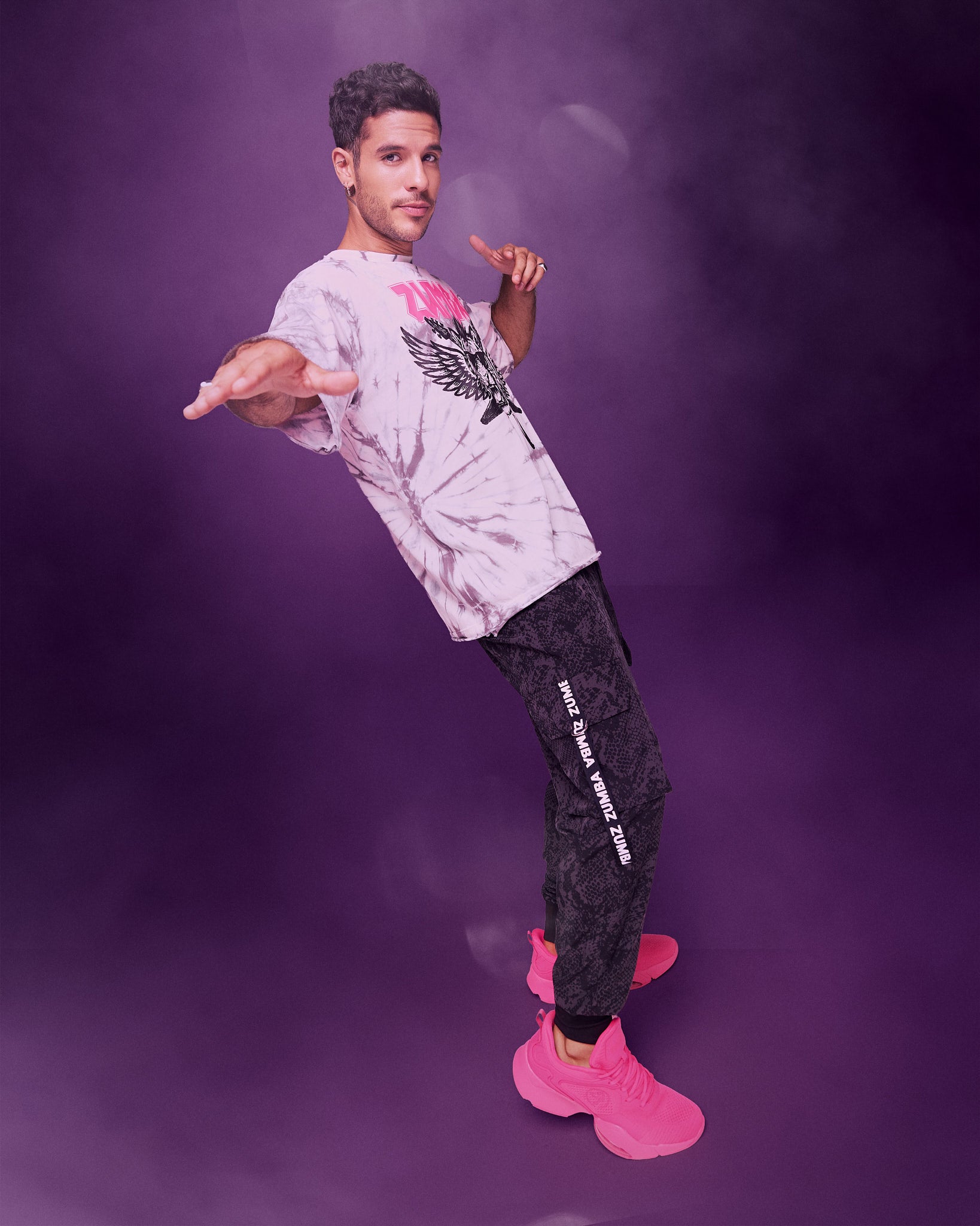 A male model posing in a studio setting with a purple smoke background. He is wearing a light white tie-dyed patterned top, black and grey cargo pants and pink shoes.
