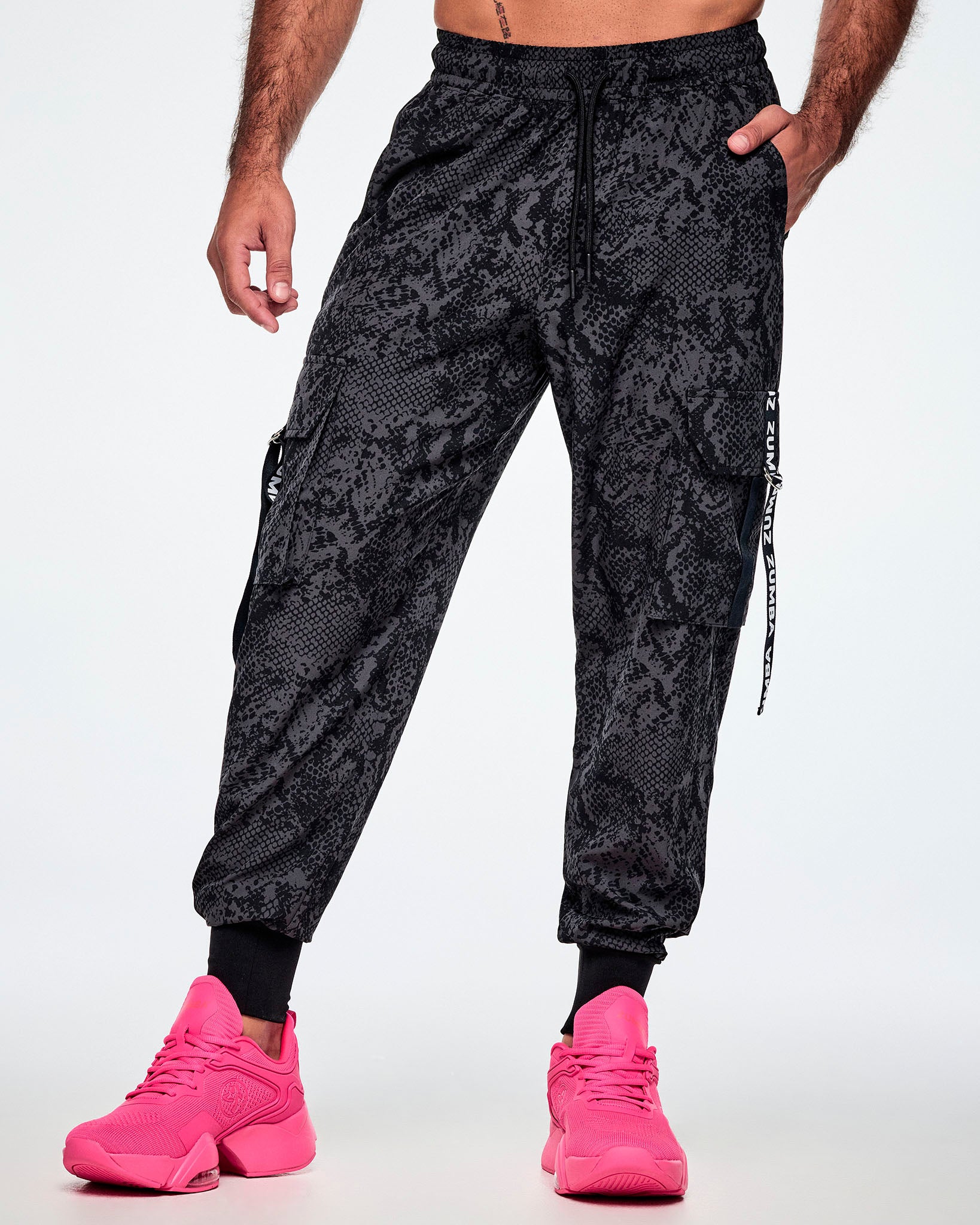 Front view of a male model wearing black and grey snake-skin patterned cargo pants with white vertical Zumba text the black straps on the cargo pockets and paired with pink shoes.