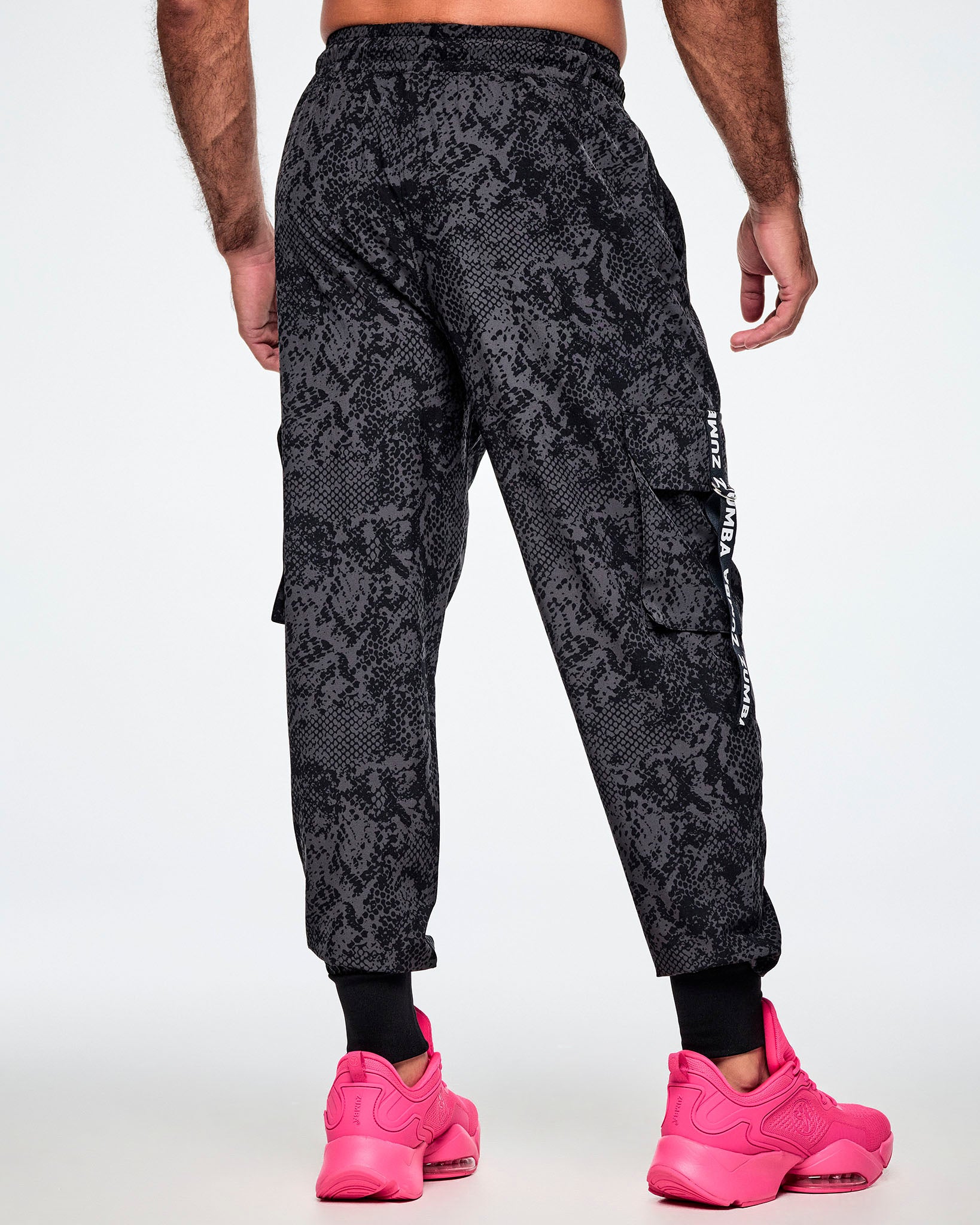 Back view of a male model wearing black and grey snake-skin patterned cargo pants with white vertical Zumba text the black straps on the cargo pockets and showing the black leg cuffs while paired with pink shoes.