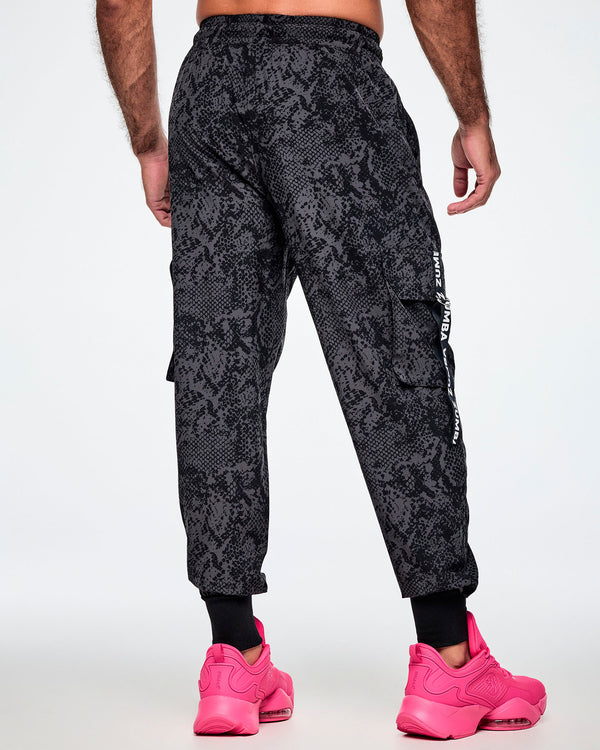 Back view of a male model wearing black and grey snake-skin patterned cargo pants with white vertical Zumba text the black straps on the cargo pockets and showing the black leg cuffs while paired with pink shoes.