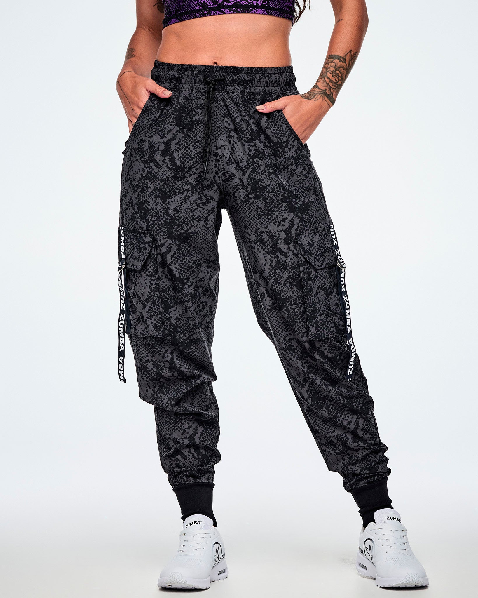 Front view of a female model, with both hands in her side pockets, wearing black and grey snake-skin patterned cargo pants with white vertical Zumba text the black straps on the cargo pockets and paired with white shoes.