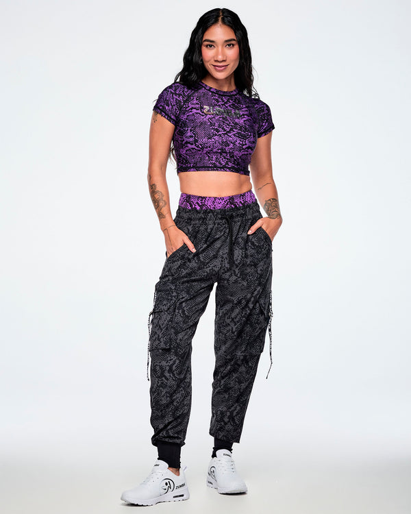 A female model wearing a purple snake-skin crop top and matching snake-skin grey cargo pants, paired with white shoes.