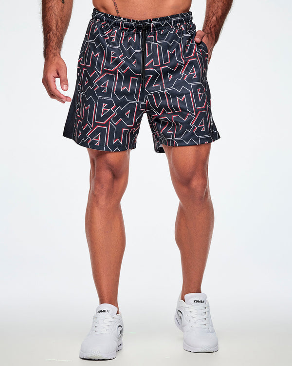 Male model with a hand in his pocket of Zumba mesh shorts featuring a bold black, white, and red ZUMBA text pattern, paired with white shoes.