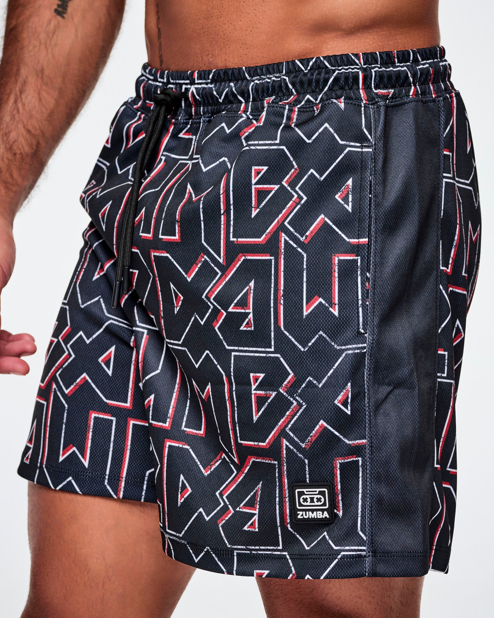 Close-up view of a male model wearing mesh shorts featuring a bold black, white, and red ZUMBA text pattern, a black stripe down the side and a ZUMBA cassette tape emblem on the lower left leg.