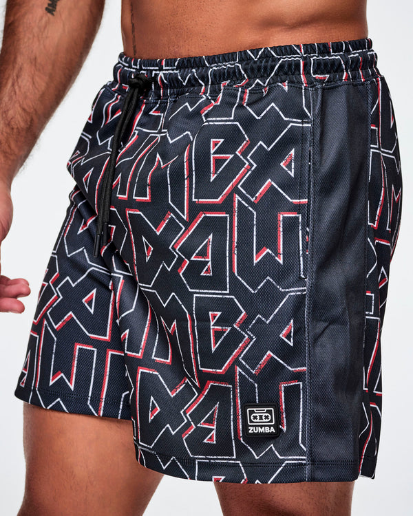 Close-up view of a male model wearing mesh shorts featuring a bold black, white, and red ZUMBA text pattern, a black stripe down the side and a ZUMBA cassette tape emblem on the lower left leg.