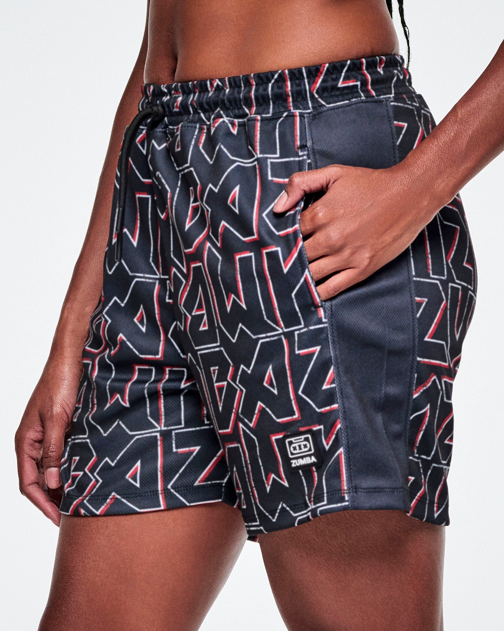 Close-up view of a female model wearing mesh shorts featuring a black, white, and red ZUMBA text pattern, a black stripe down the side and a ZUMBA cassette tape emblem on the lower left leg.