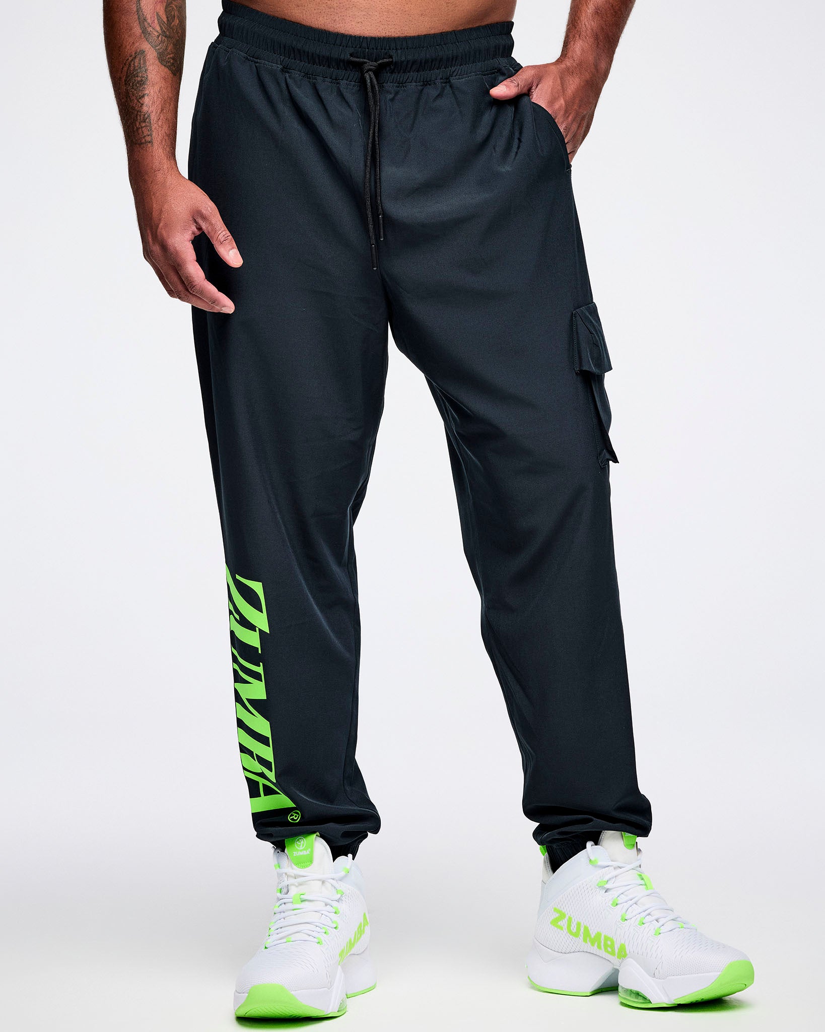 Front view of a male model with his left hand in the pocket of black cargo pants featuring green “ZUMBA” text on the right leg, paired with green-and-white shoes.