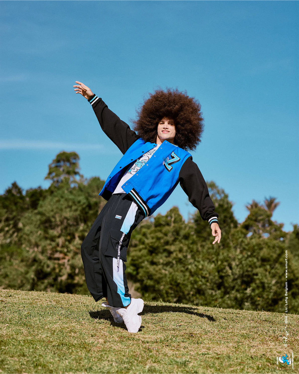 Male model leaning in a dance pose on a grassy hill, wearing the black cargo pants with light blue ombre mesh side panels and a 'ZUMBA' webbing strap, with the varsity jacket and trees behind him.