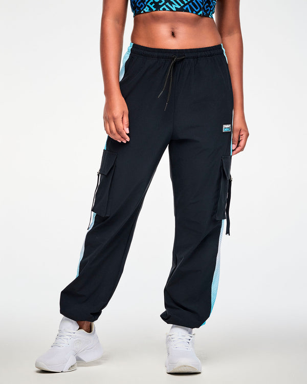 Female model in black woven cargo pants with light blue-to-white mesh side panels, a drawcord waist, elastic cuffs, and a black 'ZUMBA x THE SMURFS' patch on the left hip. Paired with white shoes.