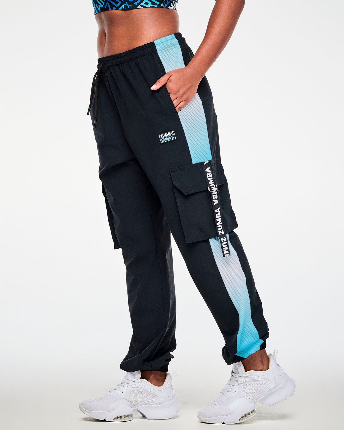 Female model with one hand in the pocket of the black cargo pants, shown in side view with the mesh side panel and a black 'ZUMBA' webbing strap on the cargo pocket.