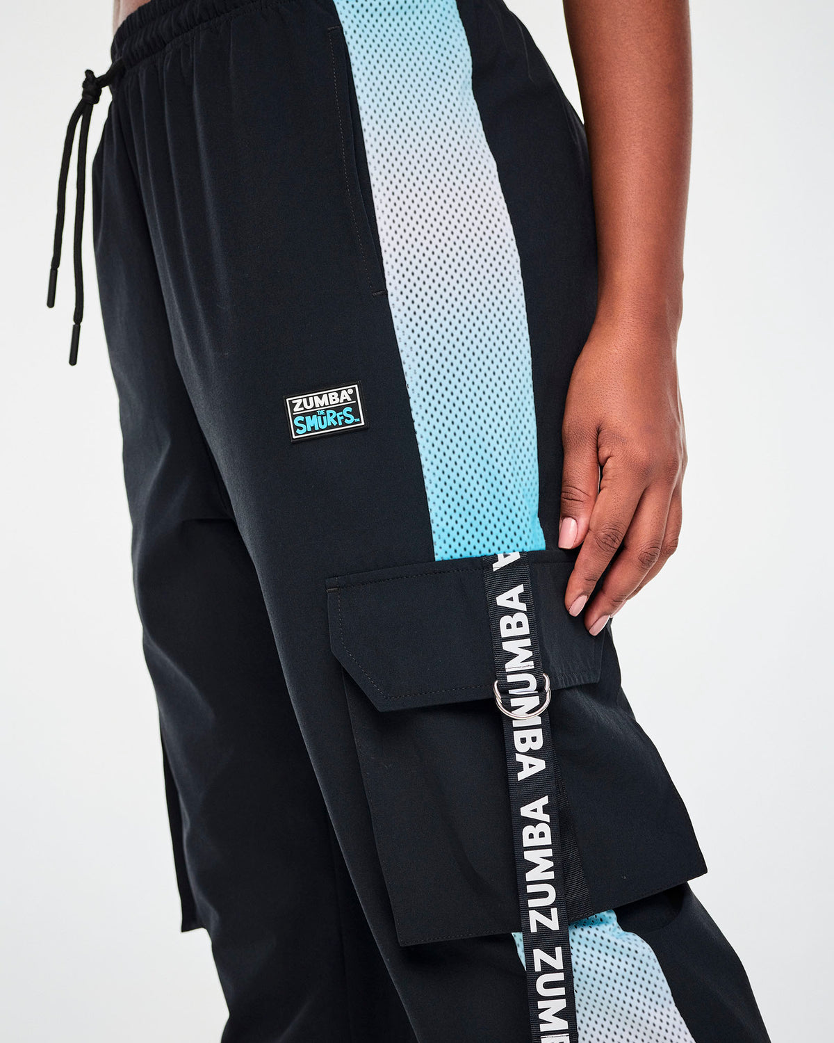 Close-up of the black cargo pants, showing the 'ZUMBA x THE SMURFS' patch, the drawcord waist, the ombre blue mesh side panel, and a 'ZUMBA' webbing strap looped through a silver D-ring on the cargo pocket.