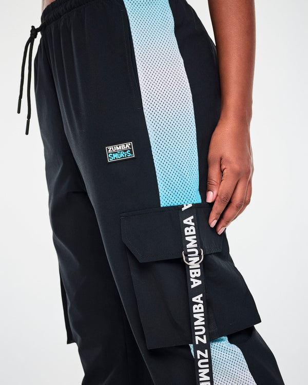 Close-up of the black cargo pants, showing the 'ZUMBA x THE SMURFS' patch, the drawcord waist, the ombre blue mesh side panel, and a 'ZUMBA' webbing strap looped through a silver D-ring on the cargo pocket.
