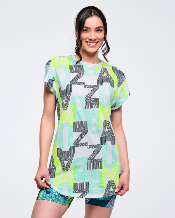 Longline cap-sleeve top with all-over “ZUMBA” letter print in aqua, lime, and gray, worn by a female model with green-and-blue patterned biker shorts.