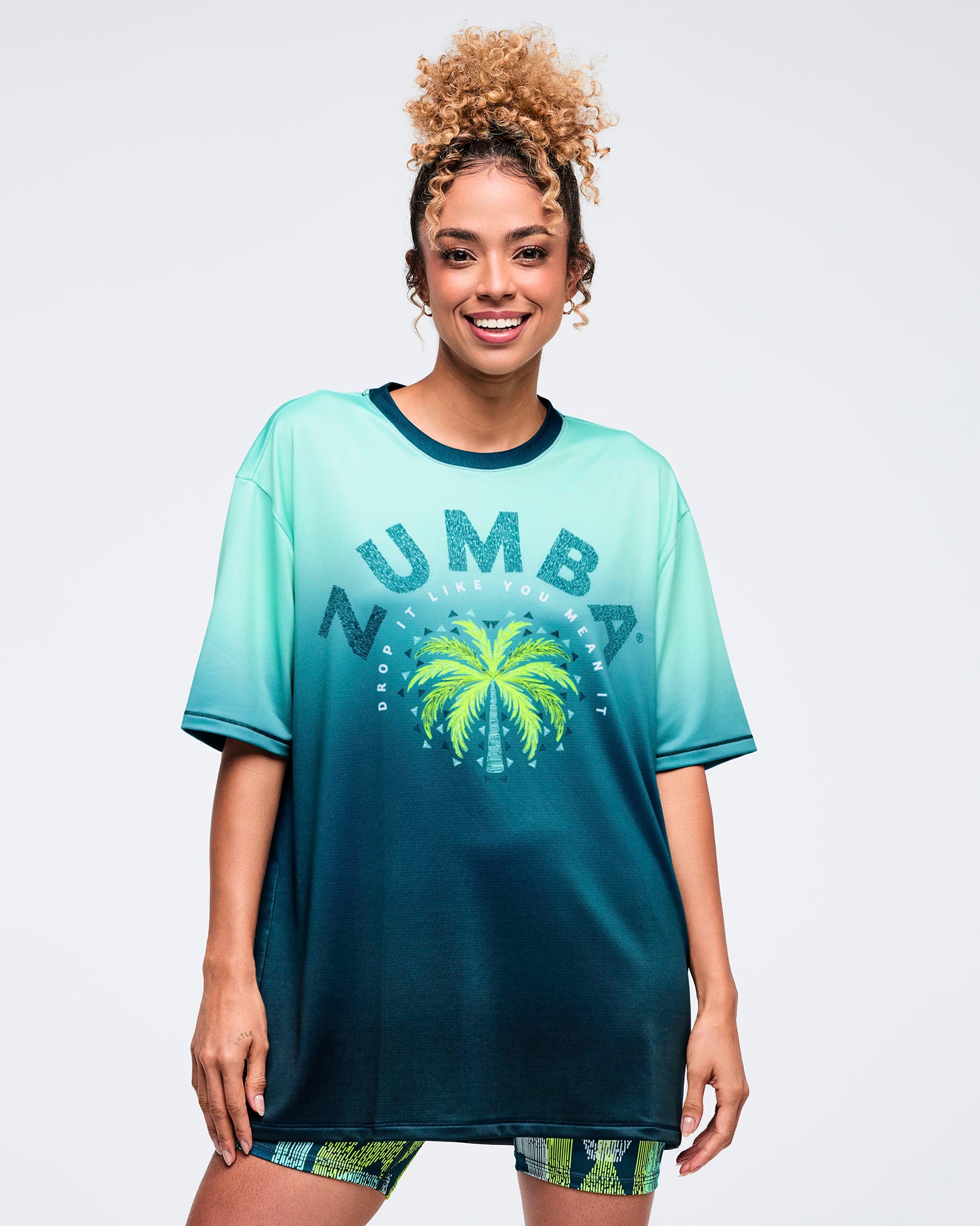Oversized ombre aqua-to-deep teal graphic T-shirt with dark crewneck, large “ZUMBA” text and palm tree print on the front, worn by a female model over green-and-teal patterned biker shorts.