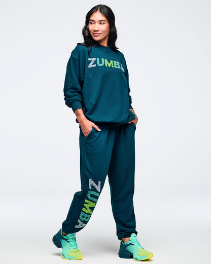 Teal crewneck sweatshirt styled with matching teal joggers featuring a vertical “ZUMBA” leg graphic, modeled by a female wearing teal-to-lime athletic sneakers on a white studio background.