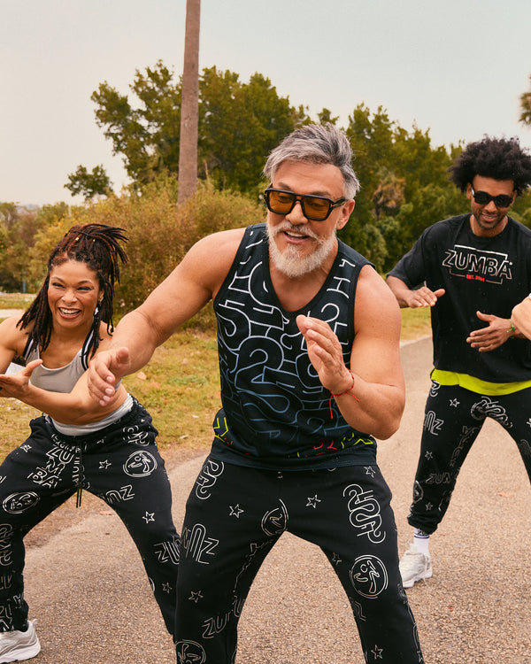 Beto, Zumba's founder, wears the fitted black tank, matching black joggers with silver Zumba graphics and white sneakers dancing on a road beside a female model in a silver sports bra and black printed joggers and a male model in a black graphic tee.