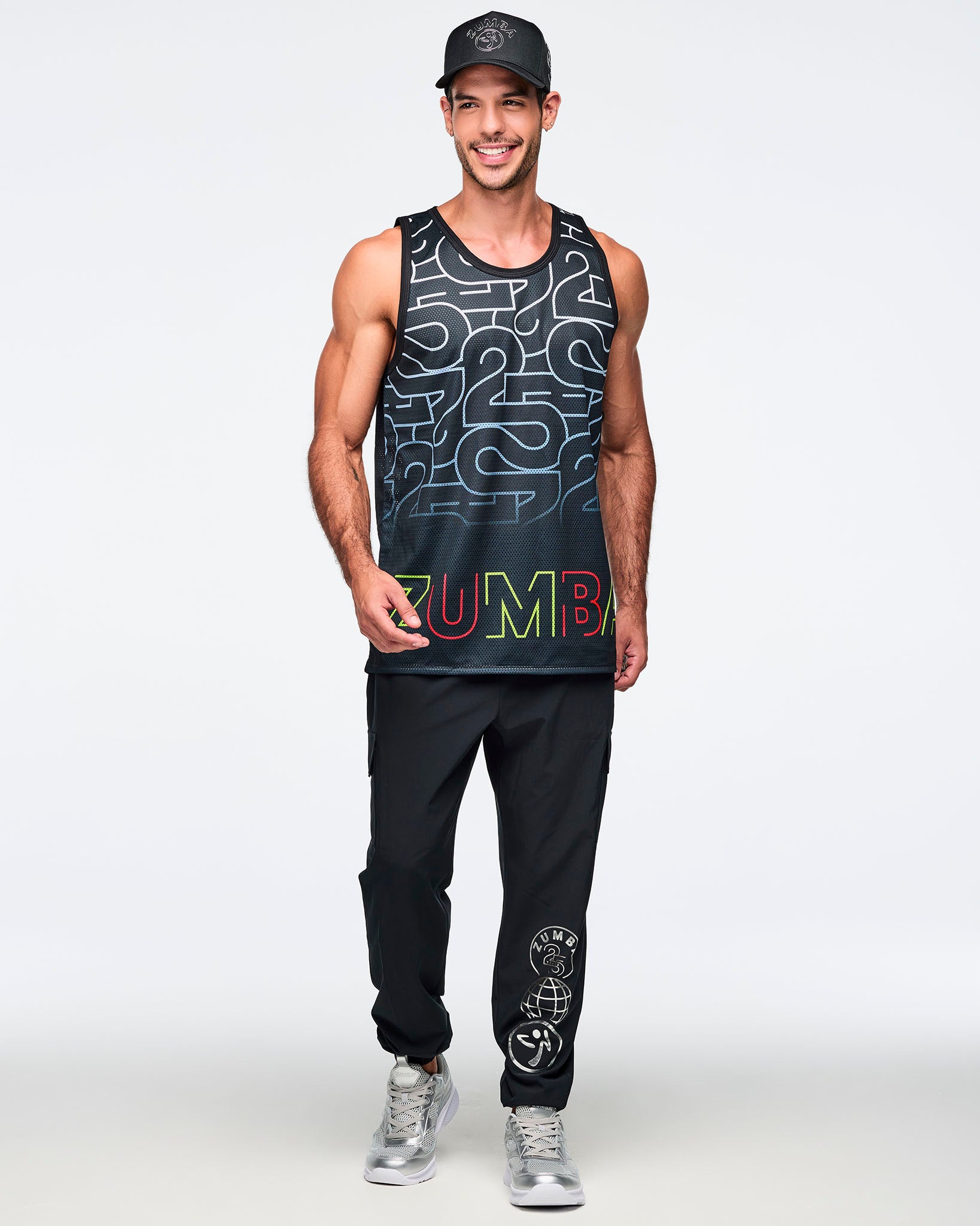 Black mesh tank with an allover silver 25 graphic and multicolor Zumba logo at the hem. A smiling male model wears the sleeveless longline tank with black jogger pants, with silver graphics on the left leg, silver sneakers, and a black baseball cap against a light gray background.